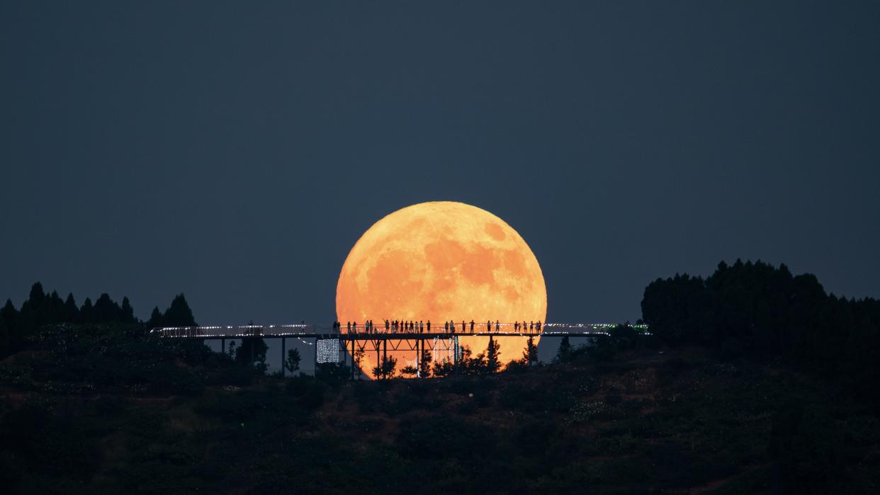 Tiny outlines of people standing on the Longquan Mountain Observation Deck from a distance against a spectacular view of a large supermoon as it rises in the night sky.