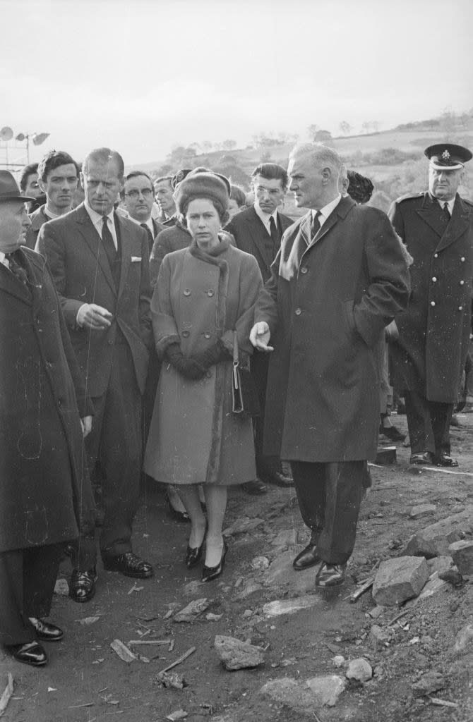 the Queen in a coat and hat standing with a group of men in the devastation