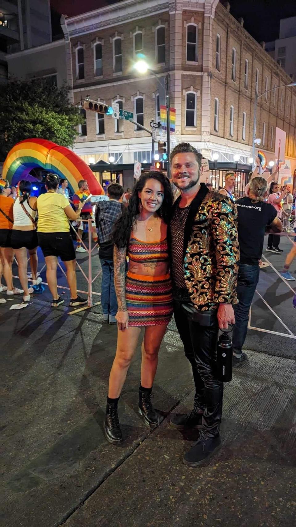 Two individuals stand at an outdoor event, one wearing a patterned short dress and boots, the other in a gold-patterned jacket and black pants. A colorful arch is in the background