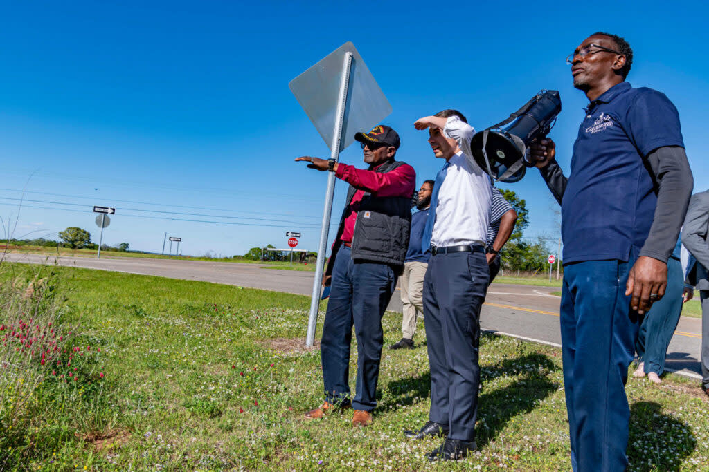 A man in a red shit and a baseball cap pointing at something; a man in a shirt and tie covering his eyes to see it; a man holding a bullhorn