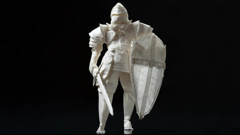 An origami knight made of rice paper with a shield and sword stands in front of a pure-black background.