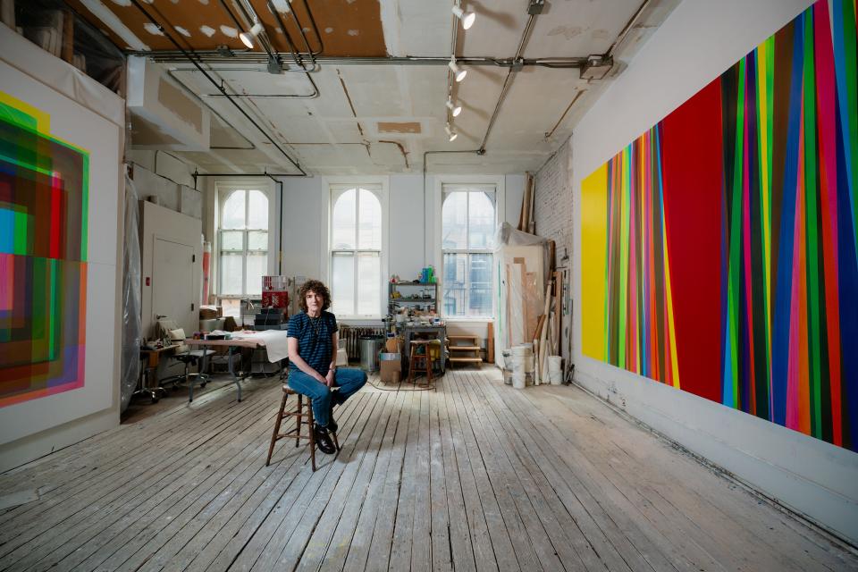 Betsy Kaufman sits in her industrial studio space with rainbow colored paintings on the wall.