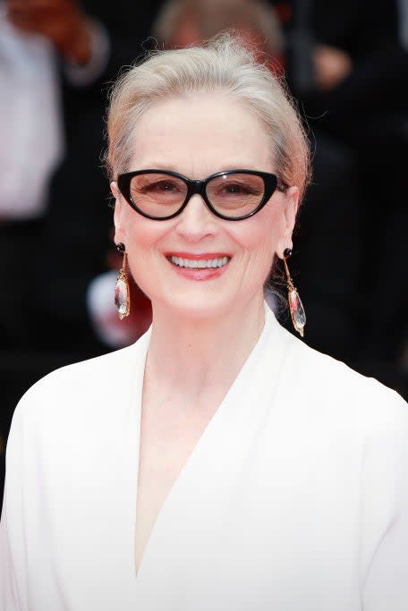 Meryl Streep on the red carpet, wearing a white elegant gown and stylish glasses, smiling at the camera