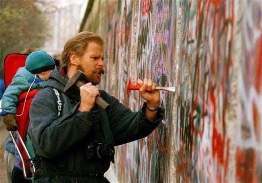 A sight-seeing West Berliner carries his baby and uses a hammer and chisel to carve out a piece of the Berlin Wall, Nov. 14, 1989. Many souvenir hunters come to the Berlin to break pieces off the crumbling wall.