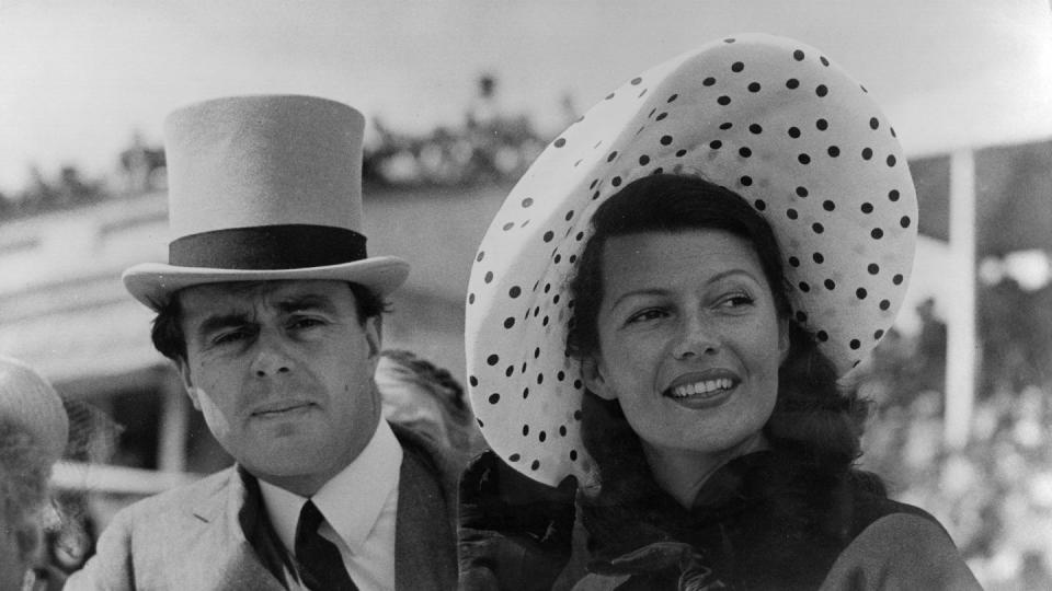 aly khan and rita hayworth stand behind a wall, he wears a suit with a tie and top hat, she wears a polka dot hat with a coat and gloves