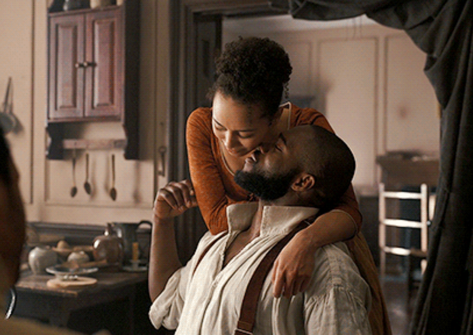 A woman embraces a man from behind, both dressed in period attire, in a tender moment indoors