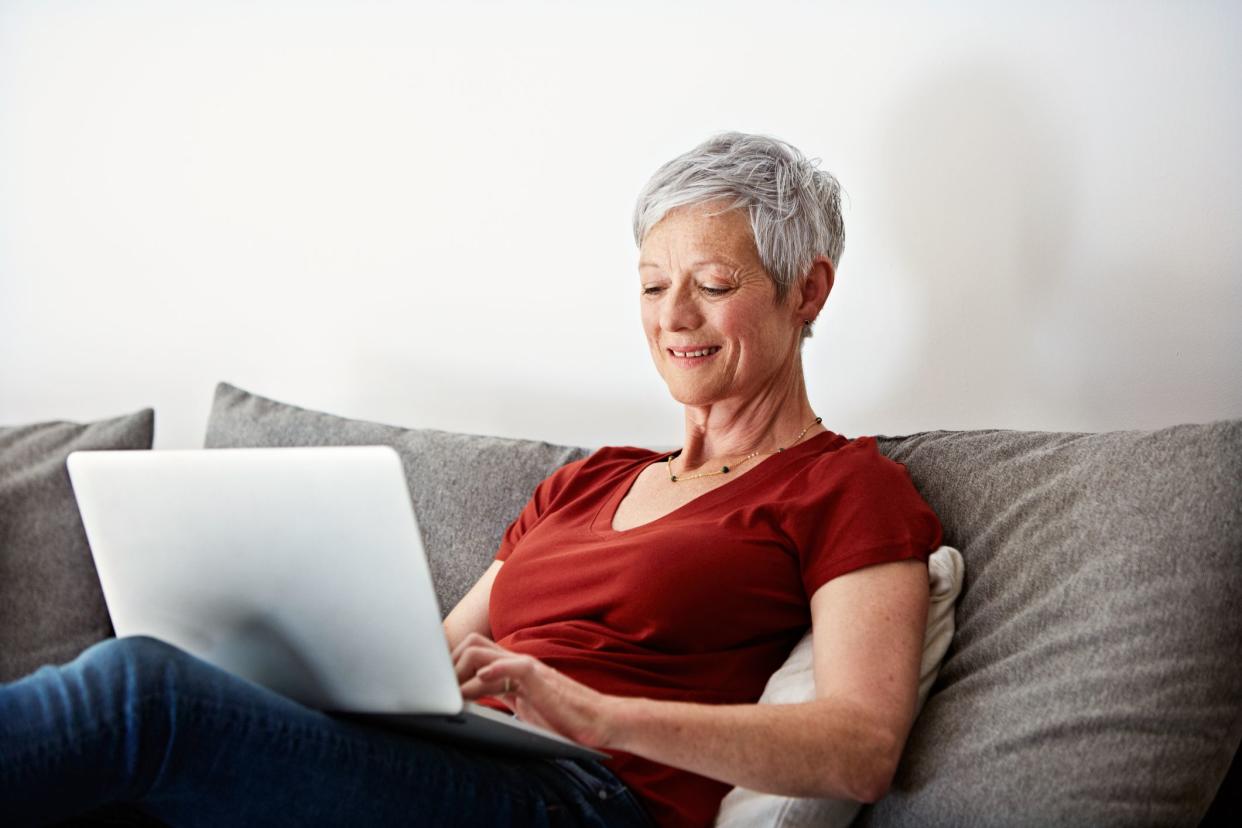 Satisfied senior woman sitting comfortably on couch while working on her laptop, grey couch, wearing casual clothing, with a white wall in the background