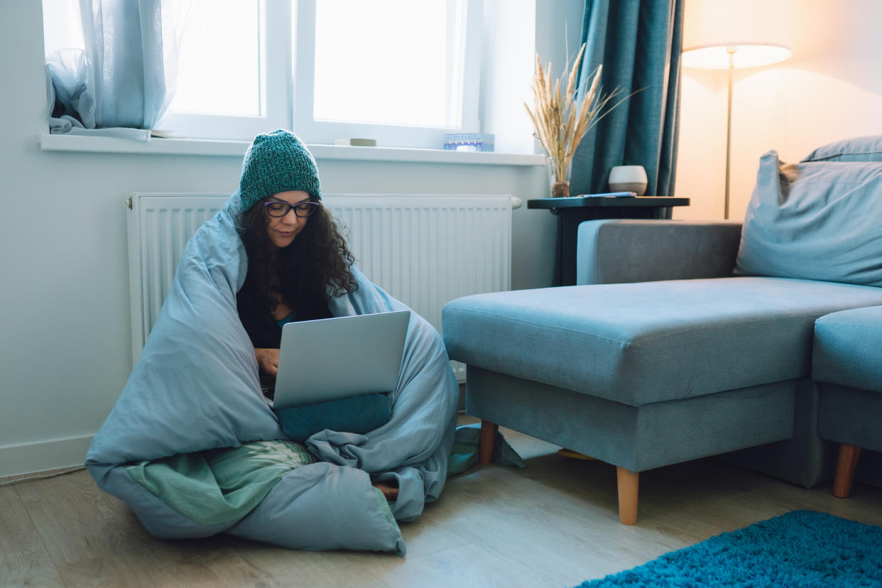 Middle age business woman work at home sitting on a floor next to heater wrapped in blanket and wearing woollen hat