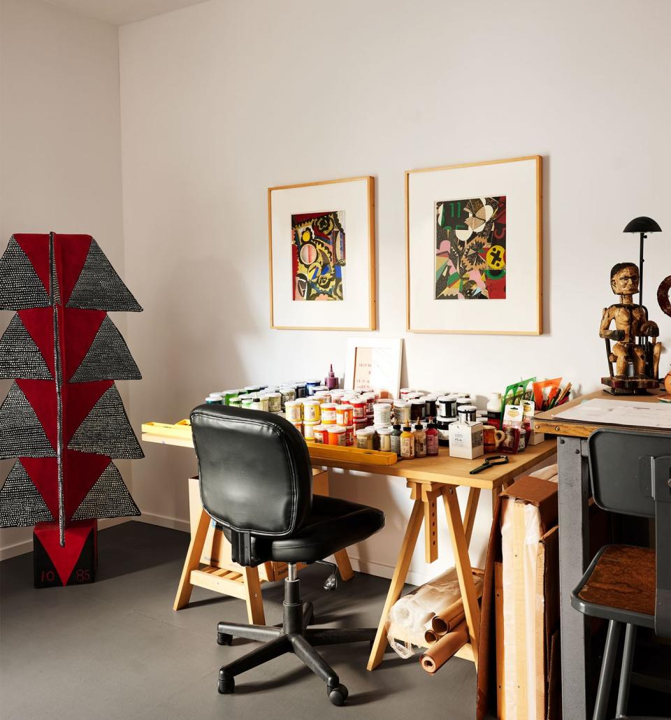 a painter's studio has a natural wood sawhorse table covered with multicolored jars of paint, a black desk chair, two framed collages above table, and a painted wood and papier mache sculpture on floor at left