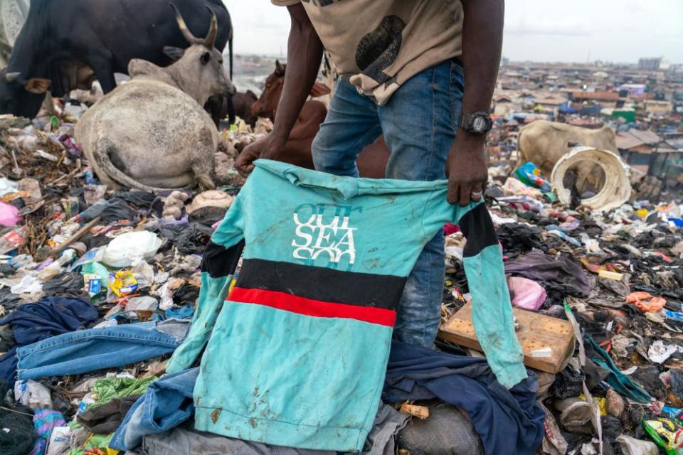 A man stands on a rubbish tip holding a dirty shirt with the slogan 'Our sea son'. Cows sit and stand on waste behind him while a shanty town can be seen in the distance