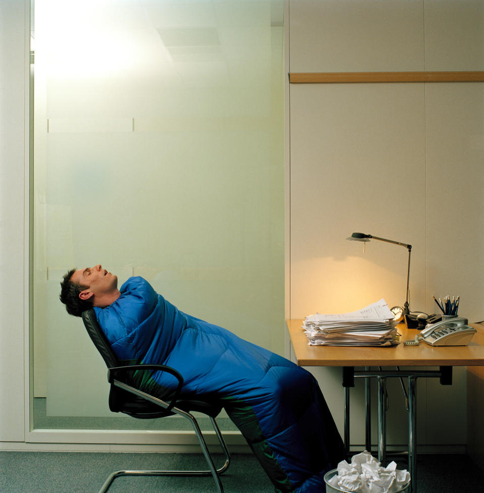 A man sleeping in an office chair while wrapped in a sleeping bag