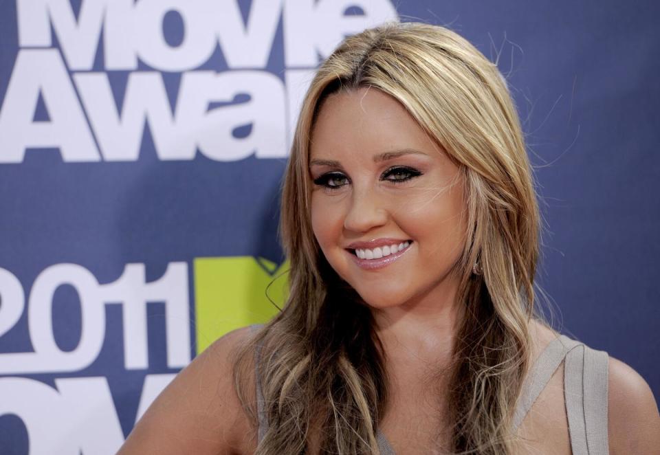 Amanda Bynes with long wavy blond hair poses in front of a blue backdrop.