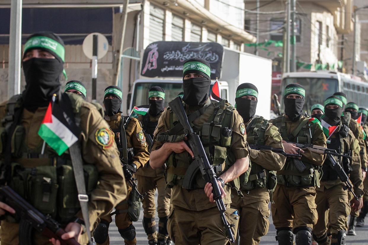 Military members hold guns and march down the street. Their faces are partially covered, and they wear beige uniforms and carry the Palestinian flag.