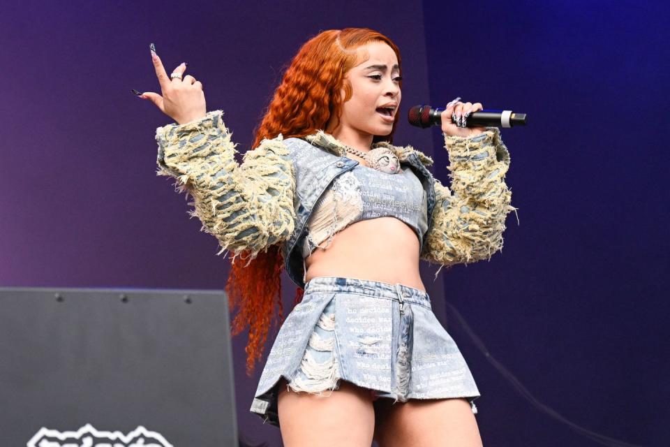 ice spice sings into a cordless microphone she holds in one hand and points the index finger of her other hand toward the sky as she stands on a stage, she wears a denim crop top and mini skirt with a matching cropped jacket