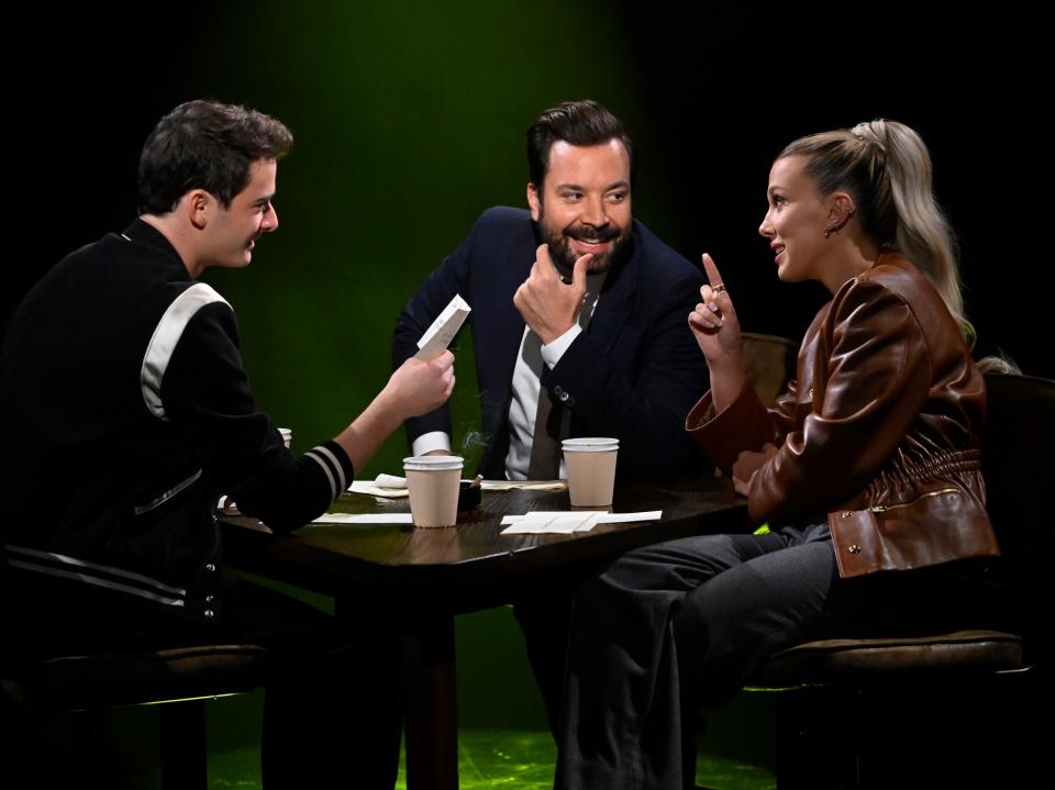 from left to right, noah schnapp, jimmy fallon, and millie bobby brown sitting at a triangular table, looking at each other with engaged, comedic expressions on their faces. schnapp is holding up a piece of paper, and fallon and brown are looking at each other