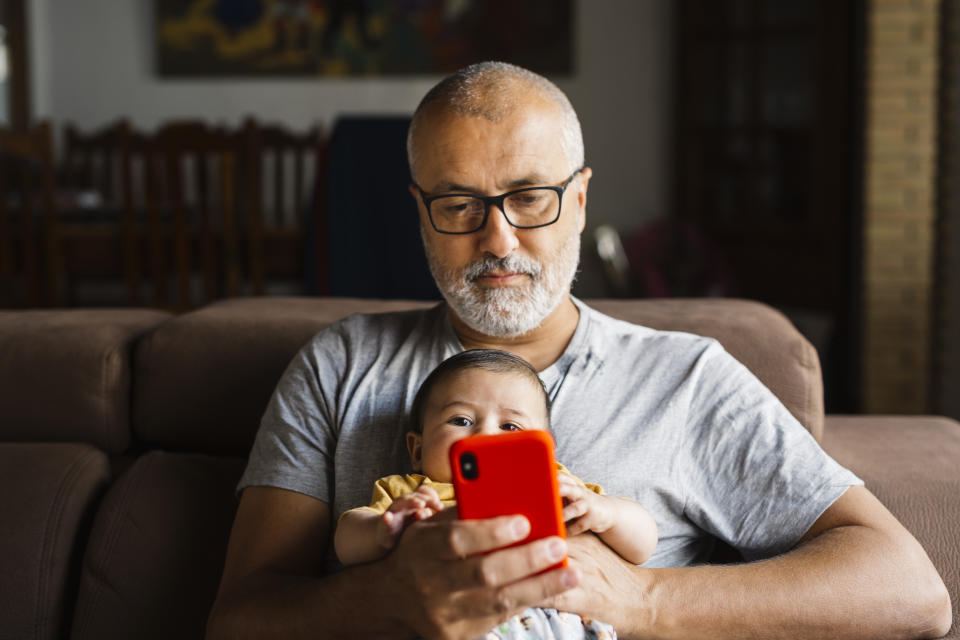 cost White haired middle aged man sitting on a sofa with his baby son while watching a red smartphone