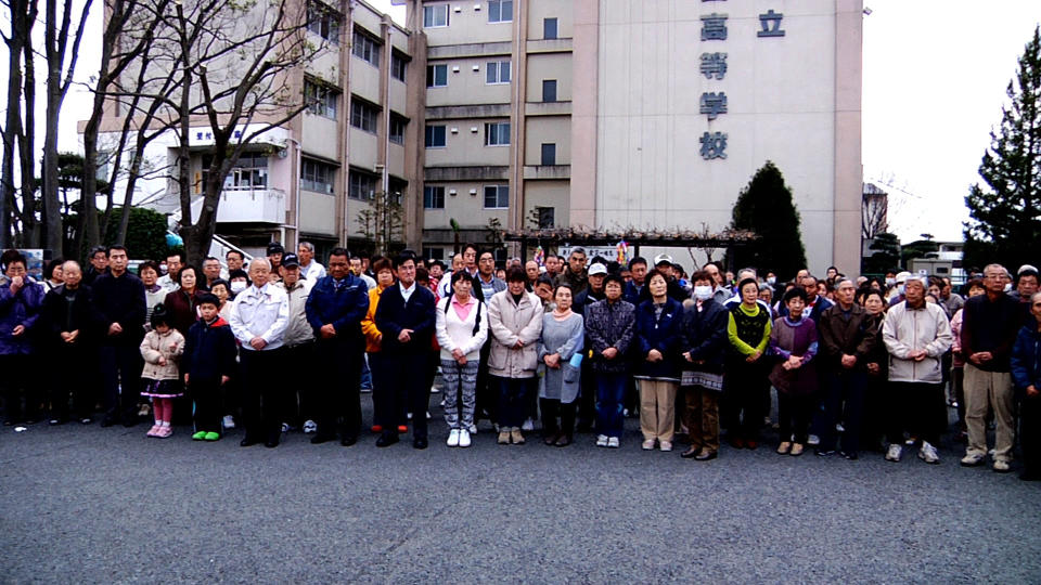 In this image made from promotional footage for the film "Nuclear Nation" released by the 2012 Documentary Japan, Big River Films, Futaba mayor Katsutaka Idogawa, sixth from left in white jacket, poses with evacuees from Futaba town in front of their shelter, the abandoned Kisai high school, in Kazo, Saitama prefecture, near Tokyo, on March 11, 2012, a year after the tsunami and earthquake hit northern Japan. The film "Nuclear Nation," directed by Atsushi Funahashi, documented a story of the residents of Futaba, the town where the tsunami crippled Fukushima Dai-ichi nuclear power plant is located. The March 2011 catastrophe in Japan has set off a flurry of independent films telling the stories of regular people who became overnight victims, stories the creators feel are being ignored by mainstream media and often silenced by the authorities. Nearly two years after the quake and tsunami disaster, the films are an attempt by the creative minds of Japan’s movie industry not only to confront the horrors of the worst nuclear disaster since Chernobyl, but also as a legacy and to empower the victims by telling their story for international audiences. (AP Photo/2012 Documentary Japan, Big River Films) CREDIT MANDATORY, EDITORIAL USE ONLY