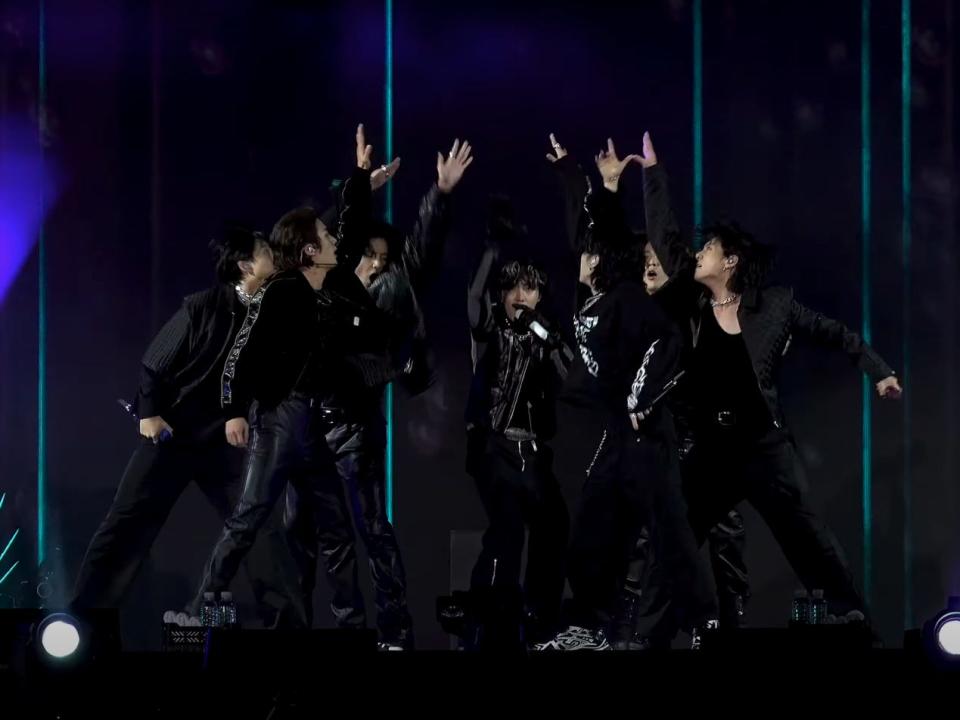 the seven members of bts, all wearing all-black outfits, standing on stage. they're in a huddle, with their arms extended upwards after placing them in the middle and extending them