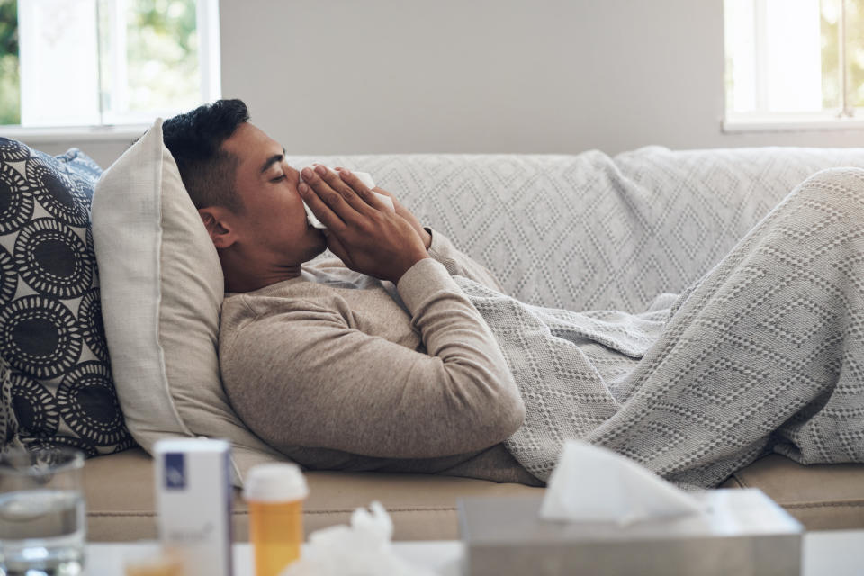 A person is lying on a couch under a blanket, using a tissue. There is medication and a glass of water on the table nearby