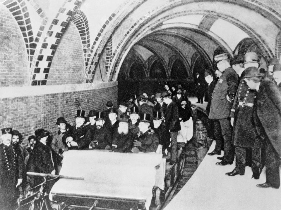 A black and white photo of the first underground subway in NYC. The cars look like roller coasters and are filled with financiers.