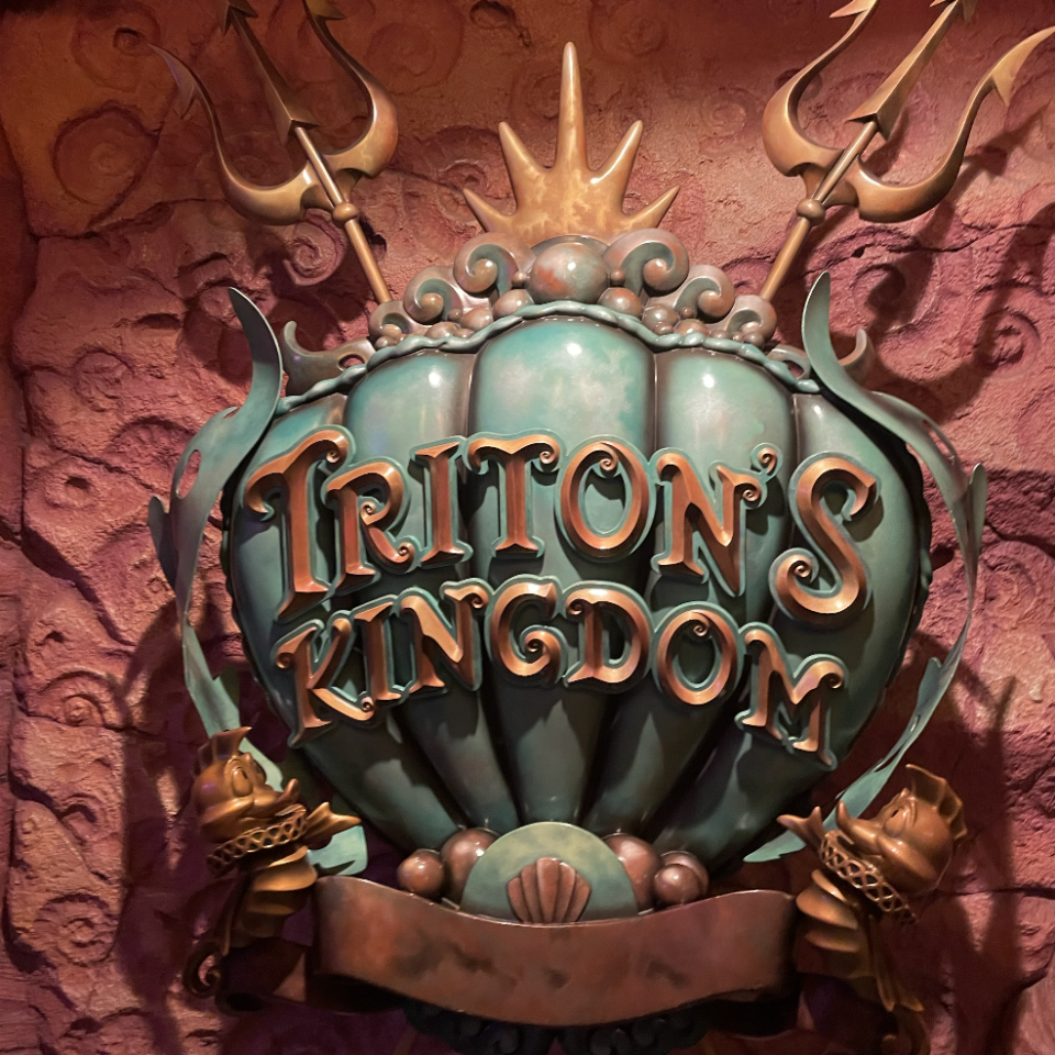 Sign for Triton's Kingdom with decorative tridents and seashells against a textured background giving an underwater feel