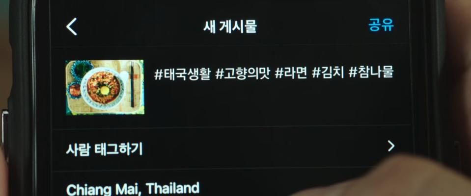a man's thumb on the screen of a cell phone, posting an image on instagram. the image, small and towards the top left of the screen, shows ramen plated with an egg, green onions, and chamnamul and kimchi on the side. the post has several hashtags in korean that read "thailand life," "ramyeon," "kimchi," and "chamnamul," and the location is tagged as chiang mai, thailand