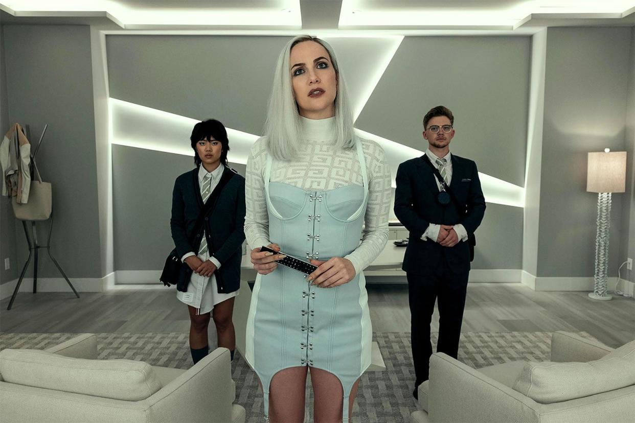 a still from The Fall of the House of Usher with three people standing in a minimalist room: a woman in the middle wears a corseted dress with a turtleneck, and two people with serious expressions flank her. 