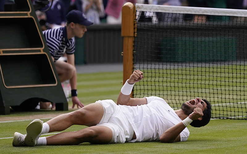 Spain's Carlos Alcaraz celebrates, lying on his back on the grass and cheering with his fists raised. Behind him can be seen the tennis net and beyond it the crowd.