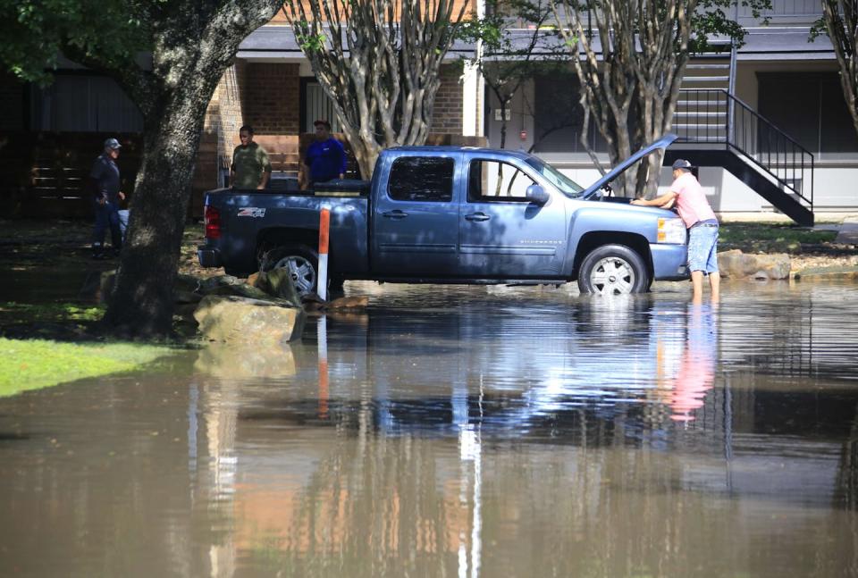 A man works on the engine of a truck while standing in floodwater over his ankles outside a home.