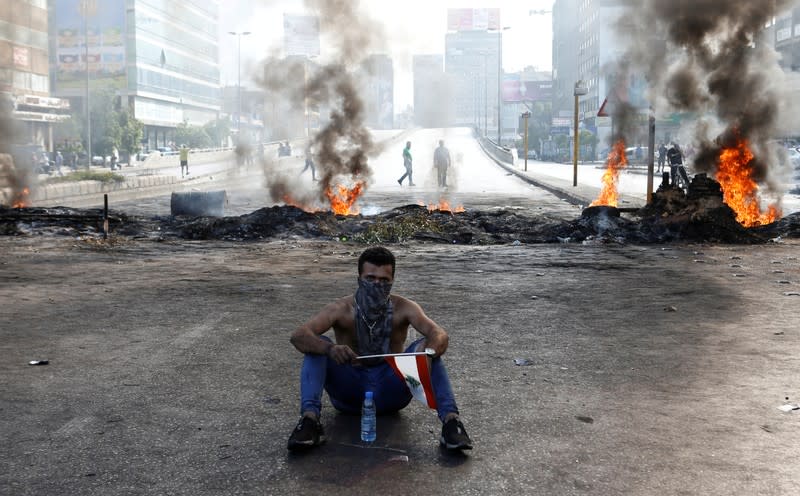 A demonstrator sits near burning tires as he holds a Lebanese flag during a protest over the deteriorating economic situation, in Dora