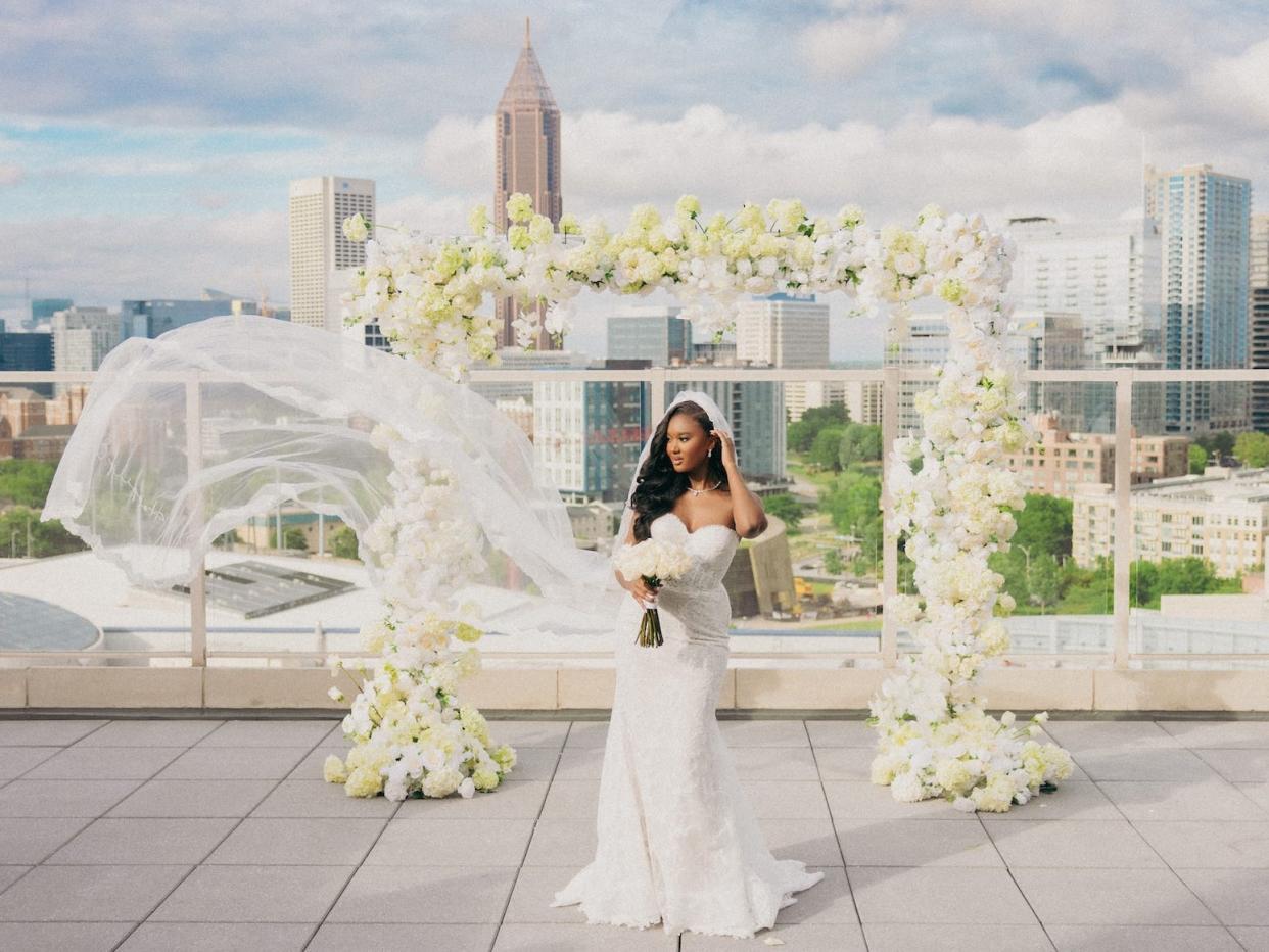 A bride stands in front of a floral archway with her veil blowing in the wind.