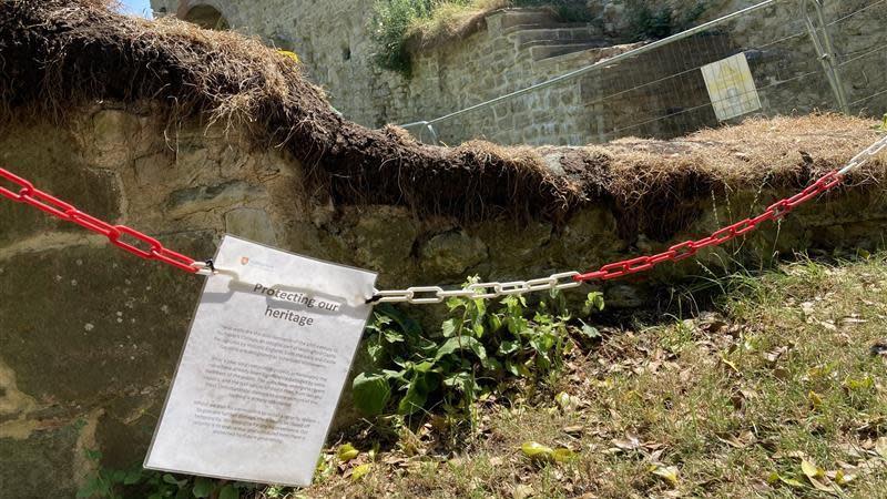 a castle wall with moss growing on it. In the foreground there is a piece of laminated paper taped to a chain which is roping off the ruins. It reads "protecting our heritage" but the rest of the page is illegible.