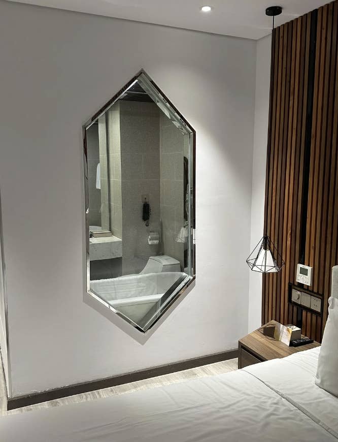 A modern hotel room features a unique hexagonal mirror, a contemporary light fixture, and a wood-paneled wall