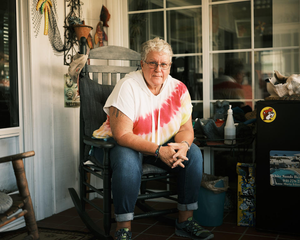 Gail Foreman sits on a rocking chair in a room with bright, natural light. She is leaning forward with her hands clasped in front and her elbows on her knees. 