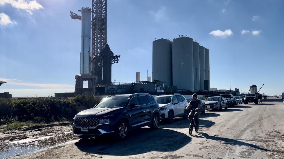 cars are parked along a dirty road  as a man on a bicycle rides parallel. An unstacked Starship rocket is seen in the background against a blue sky.