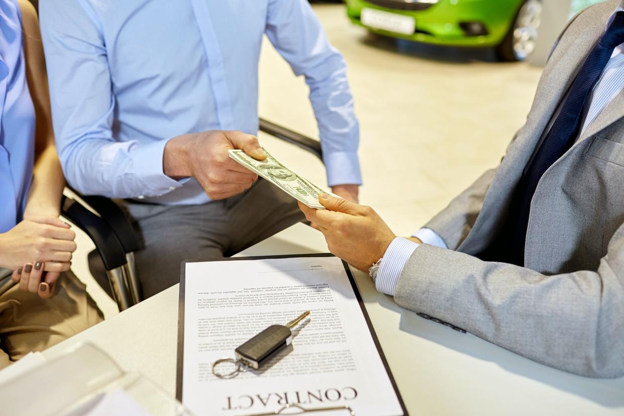 Closeup of couple giving US money to male car dealer at desk with contract and car keys with car dealership showroom in the background