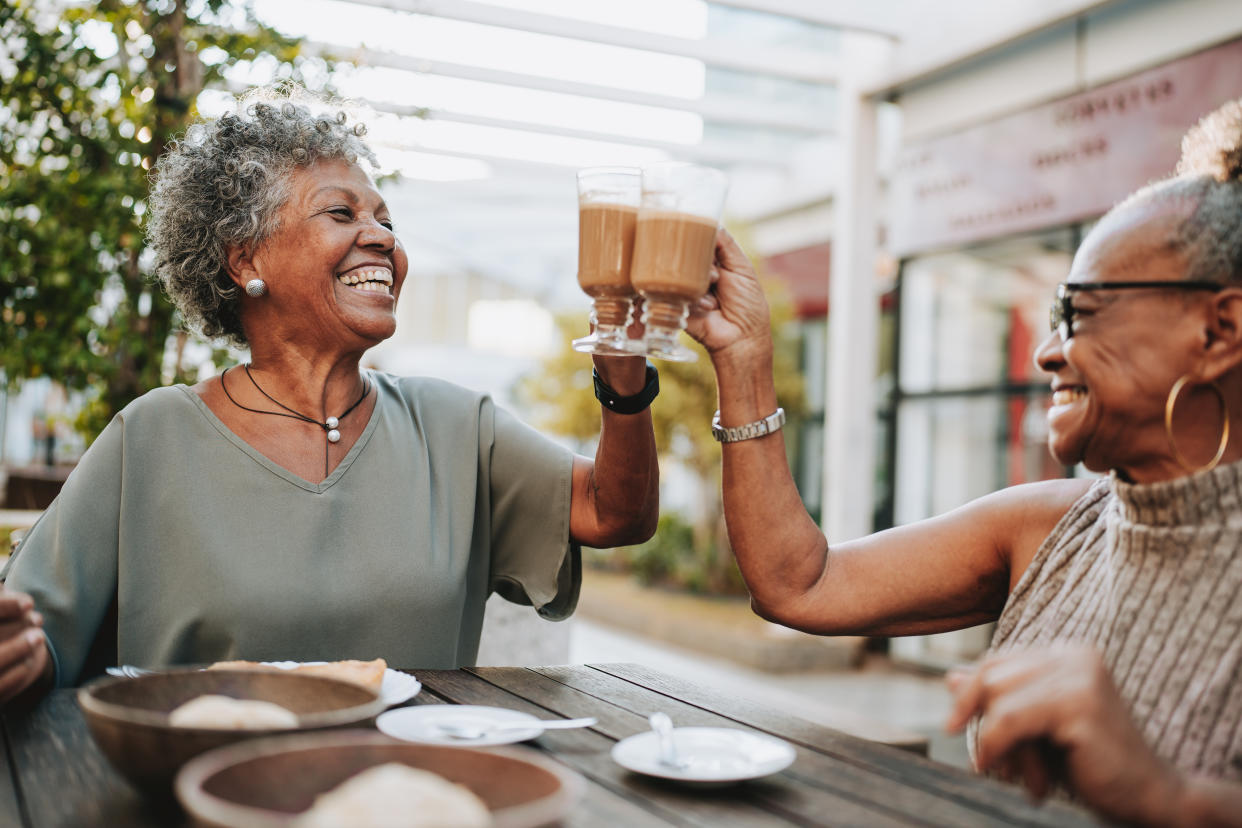 Two older women clink glasses in a toast as they sit at an outdoor table.