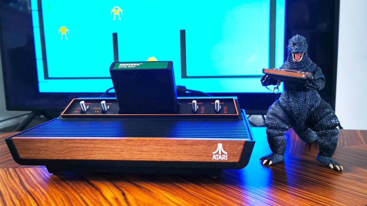  Atari 2600+ console sitting on woodgrain table with Berserk cart inserted and Godzilla toy holding tiny Atari console on right hand side. 