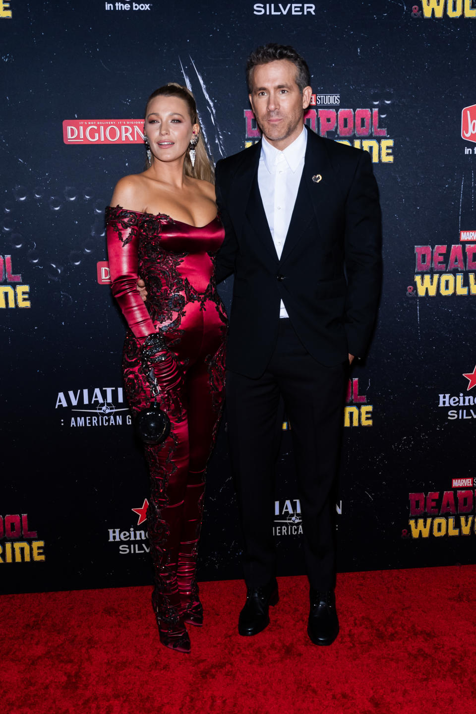 Blake Lively in a figure-hugging, off-the-shoulder red jumpsuit and Ryan Reynolds in a black suit with a white shirt pose on the red carpet of Deadpool 3