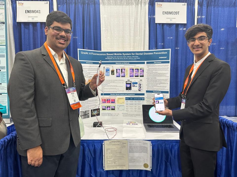 two high schoolers wearing suits and orange lanyards with conference badges stand smiling on either side of a science fair posterboard one holding a phone with an app open the other holding an electric toothbrush-like device plugged into a small black box