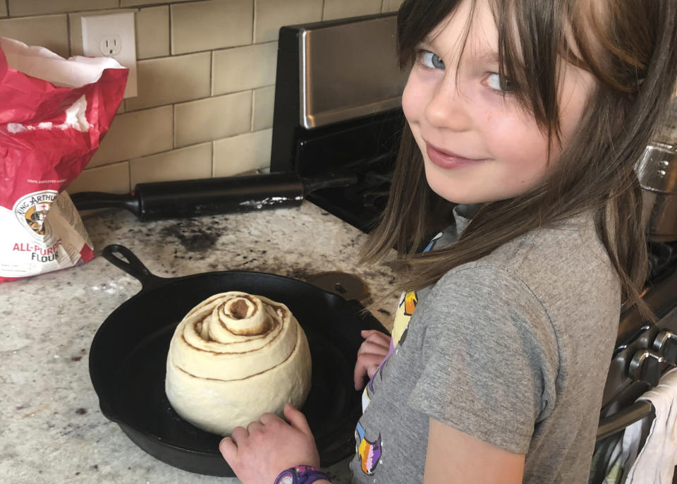 In this April 5, 2020, photo provided by Whitney Rutz, Elsa Rutz poses by a cinnamon roll at home in Portland, Ore.. Elsa's mother, Whitney, made cinnamon rolls to help raise funds for Oregon Food Bank. (Whitney Rutz via AP)