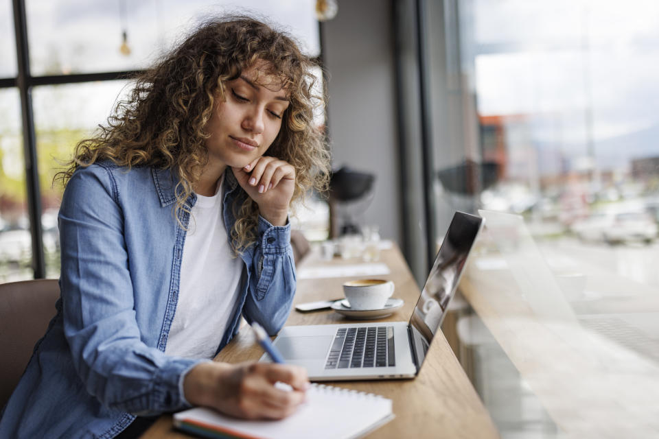 Young woman working on laptop and taking notes at a cafe, going over her mortgage renewal options