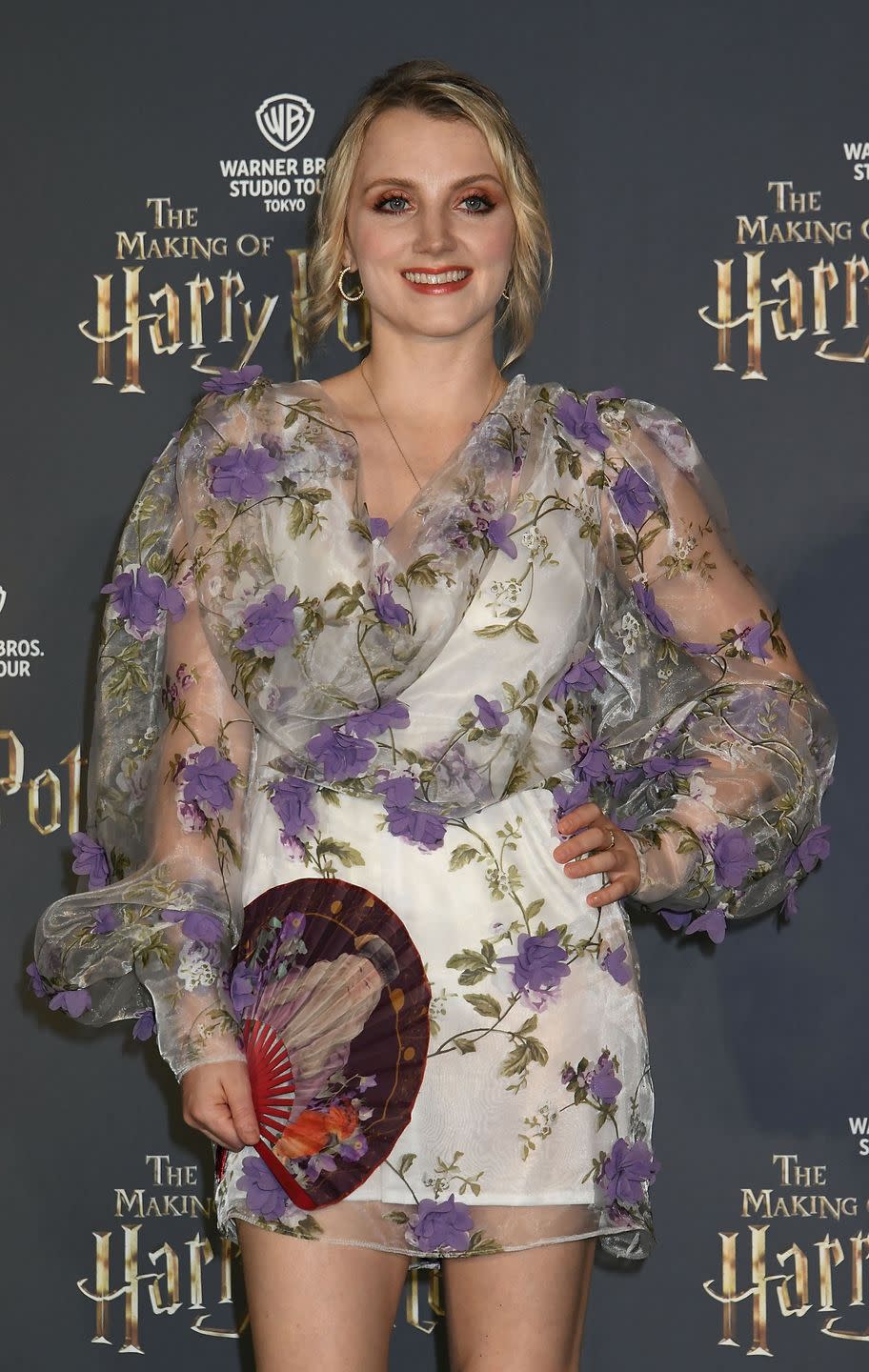 evanna lynch, a young woman stands looking at the camera with a smile and her left hand on hip, she has blonde hair worn up with curls loose framing her face and wears a white dress with purple flowers, she hold a paper fan open in her right hand