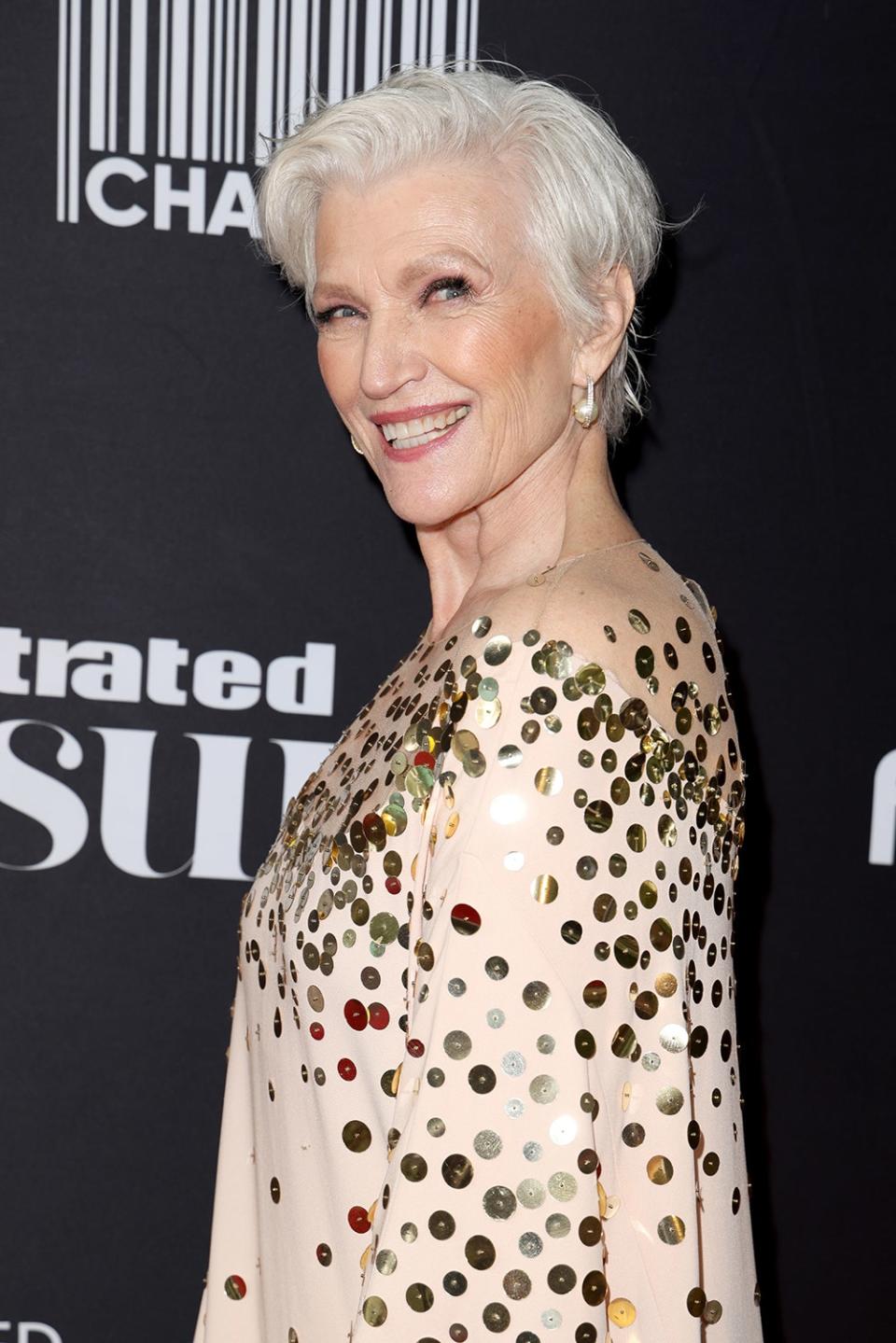 Maye Musk looking to the side and smiling in a gold and beige sparkling dress.