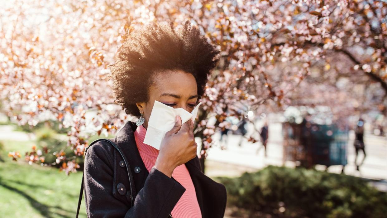  Black woman walking through a city park with blooming cherry trees in the background as she blows her nose into a tissue. 