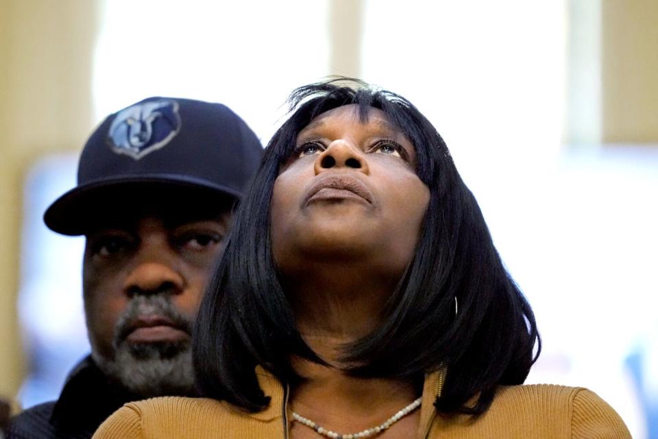 A woman with shoulder-length black hair tilting her head and gaze upward. A man in a cap stands behind her.