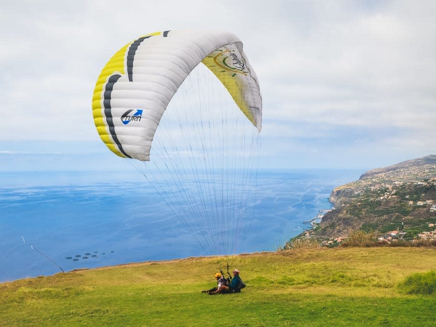 Arco da Calheta, Madeira, Portugal - Sep 16, 2019: Tandem paragliders landing on a grass on the cliffs above the Atlantic ocean. Madeiran landscape in background. Paragliding, active vacation.