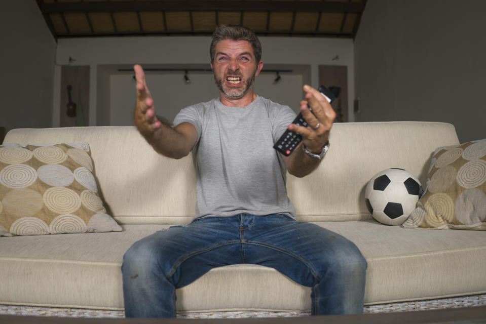 man wearing jeans and a grey t-shirt while watching a football game on television from the sofa in the living room. He is frustrated and angry his because his team is losing the game