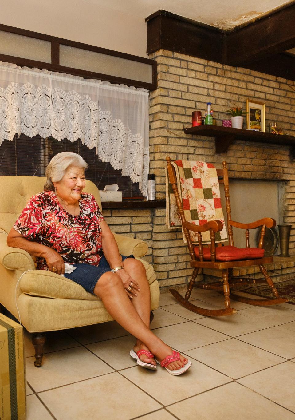 Olivia R. Hernandez sits in her favorite chair. The ceiling behind her shows damage from a leaking roof.