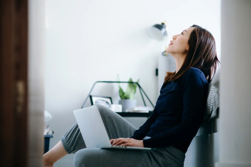 Thoughtful young Asian woman sitting on the floor in the bedroom, working from home and using laptop. Home office, remote working, freelancer, small business and lifestyle concept